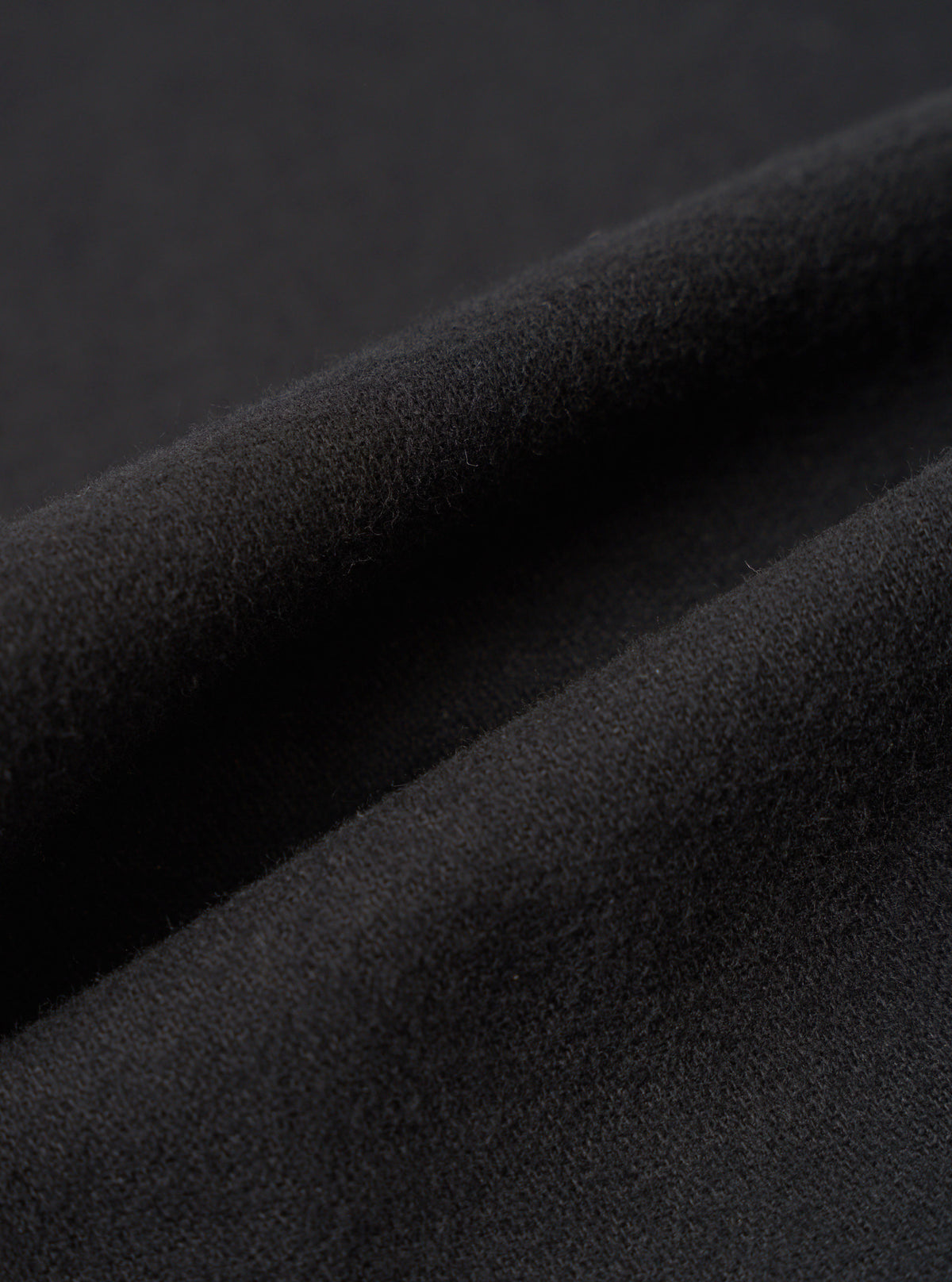 Close-up detail shot of the black brushed moleskin fabric used in the gilet. The texture is dense and softly napped with a fine, even pile visible across the surface. Deep folds in the fabric highlight its weight and drape against a dark background.