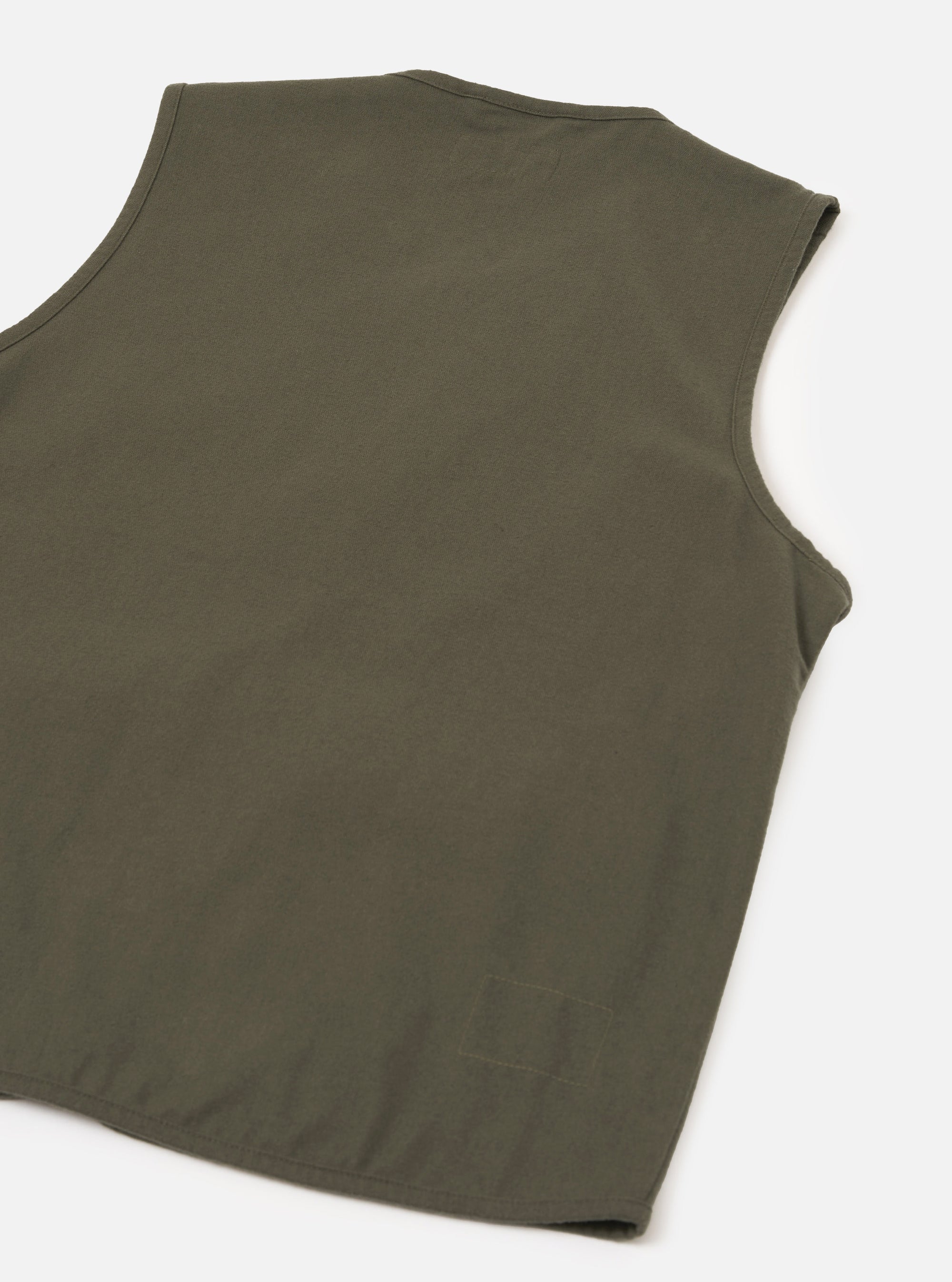 Flat lay of an olive brushed moleskin gilet on a white studio surface, shot from above at an angle. The back of the garment is shown, displaying smooth, matte fabric with a clean sleeveless silhouette, curved armholes, and a straight hem with a small side vent visible at the lower corner.