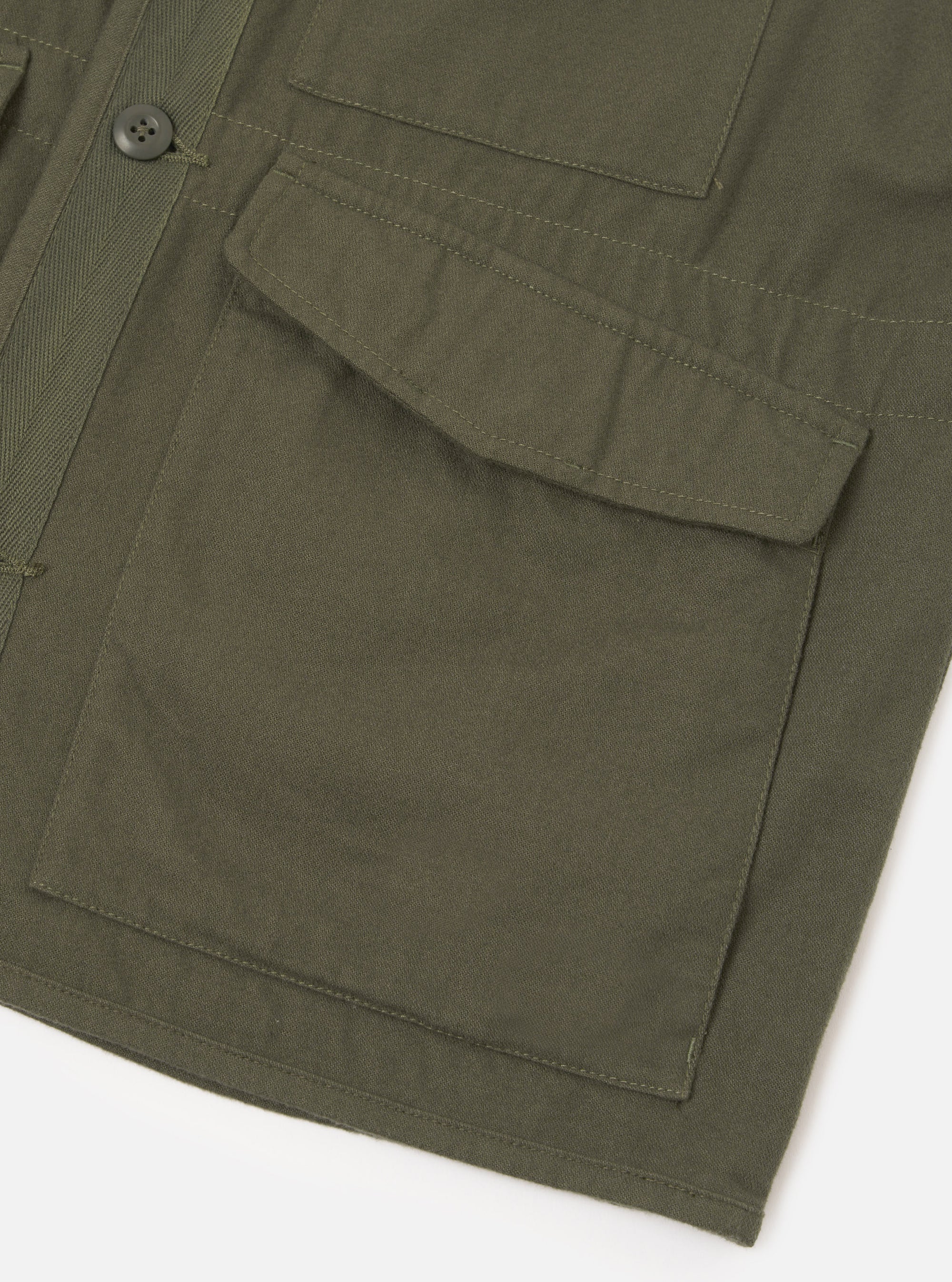 Close-up detail of an olive brushed moleskin gilet on a white studio backdrop. The shot shows two large flap patch pockets with contrast topstitching, a dark four-hole button visible at the front placket, and a herringbone-weave strap detail to the left side. The plain-weave fabric has a fine, dense texture.
