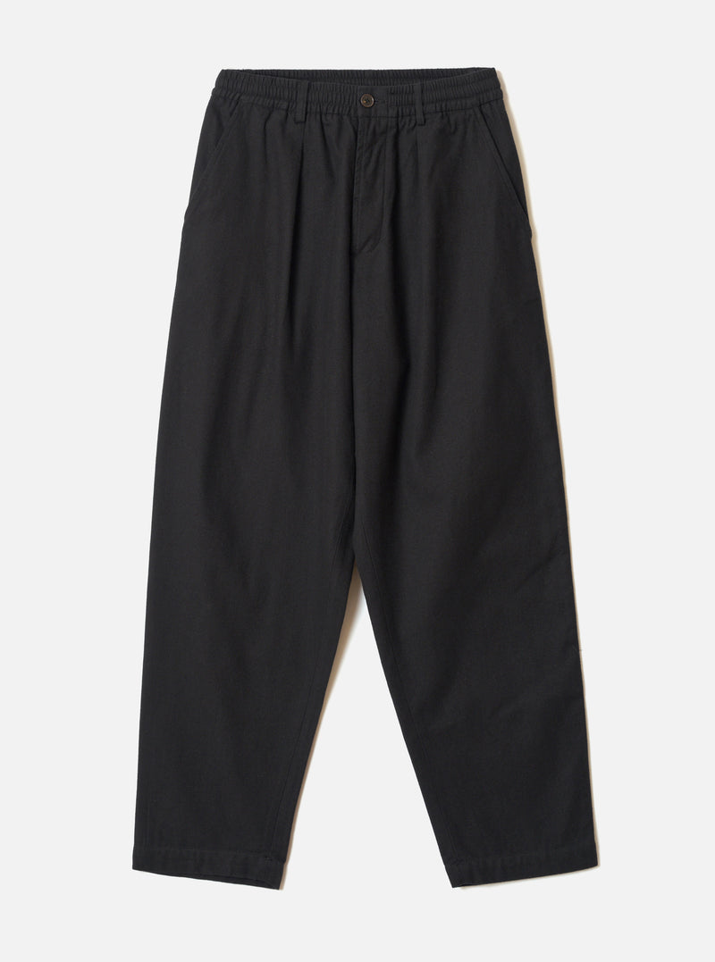 Black brushed moleskin pleated track pants laid flat on a light grey seamless backdrop. The elasticated waistband features a single dark button at the centre front. Slant side pockets sit at the hips. The wide-leg silhouette tapers slightly toward the plain, unfinished hem. Fabric has a soft, matte texture throughout.