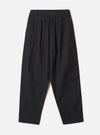 Black brushed moleskin pleated track pants laid flat on a light grey seamless backdrop. The elasticated waistband features a single dark button at the centre front. Slant side pockets sit at the hips. The wide-leg silhouette tapers slightly toward the plain, unfinished hem. Fabric has a soft, matte texture throughout.