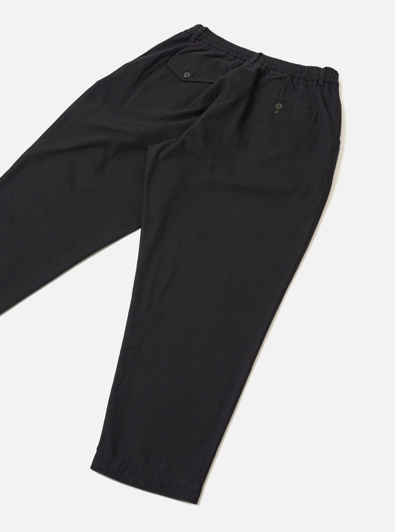 Black brushed moleskin pleated track pants laid flat on a light grey seamless surface. The elasticated waistband is visible at the top with a buttoned welt pocket on each side. A tapered leg narrows to an elasticated cuff at the ankle. The fabric has a smooth, matte finish. Shot from a diagonal angle showing both legs folded.