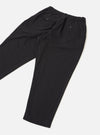 Black brushed moleskin pleated track pants laid flat on a light grey seamless surface. The elasticated waistband is visible at the top with a buttoned welt pocket on each side. A tapered leg narrows to an elasticated cuff at the ankle. The fabric has a smooth, matte finish. Shot from a diagonal angle showing both legs folded.