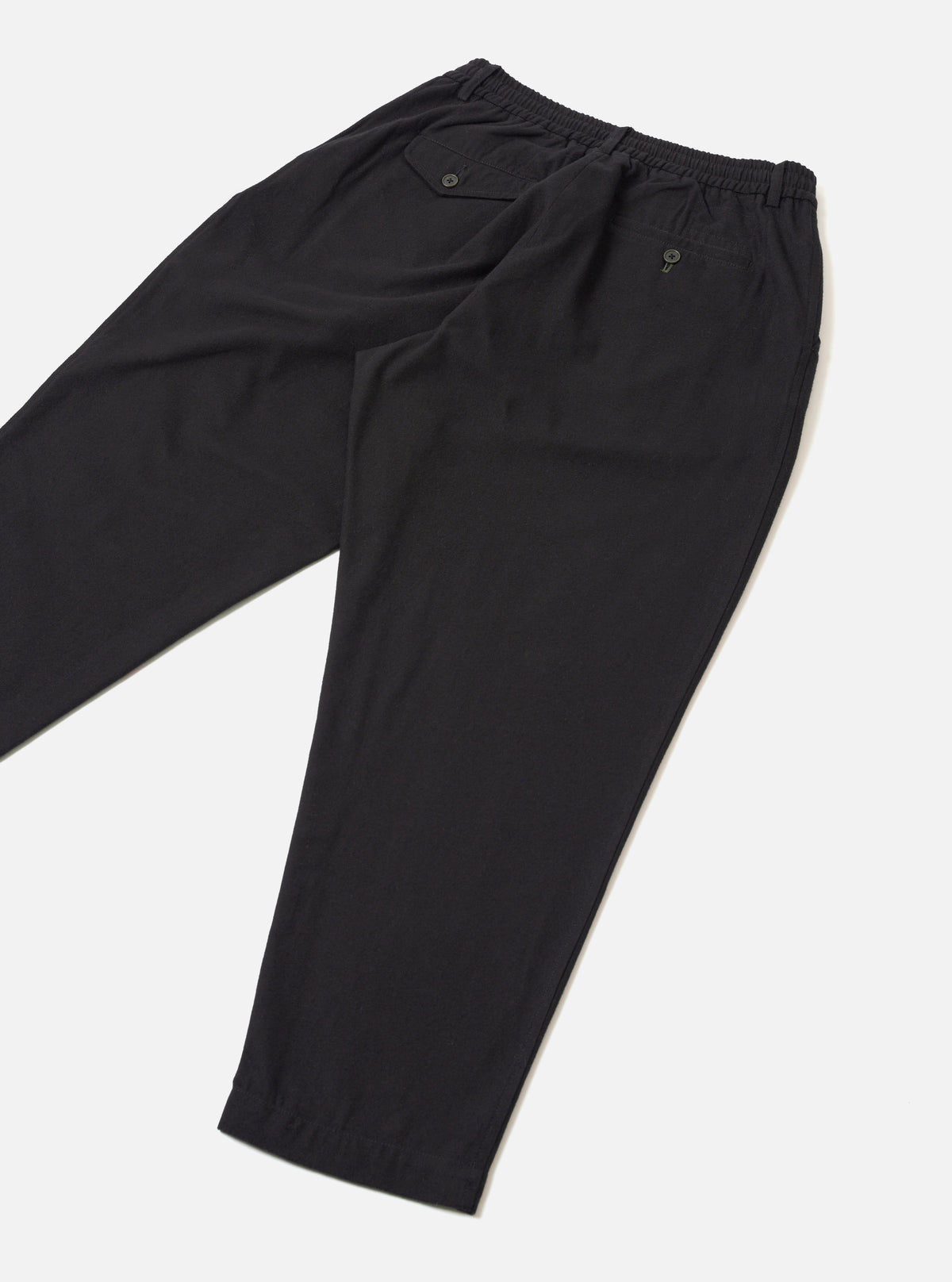 Black brushed moleskin pleated track pants laid flat on a light grey seamless surface. The elasticated waistband is visible at the top with a buttoned welt pocket on each side. A tapered leg narrows to an elasticated cuff at the ankle. The fabric has a smooth, matte finish. Shot from a diagonal angle showing both legs folded.
