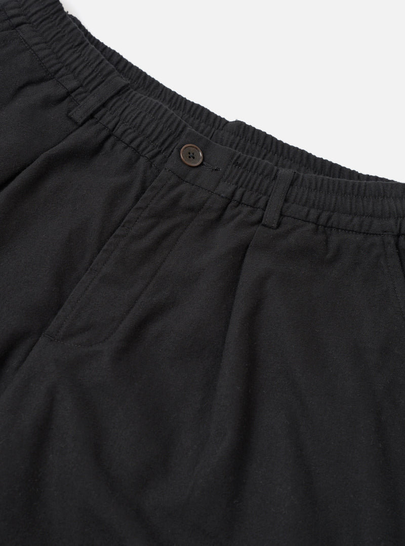 Close-up flat lay of black brushed moleskin pleated track pants on a white studio backdrop. The elasticated waistband with ruched gathering is visible at the top, alongside a single dark horn button fastening. A deep patch pocket sits at the front left, and soft fabric drape highlights the pleated construction below the waistband.