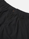 Close-up flat lay of black brushed moleskin pleated track pants on a white studio backdrop. The elasticated waistband with ruched gathering is visible at the top, alongside a single dark horn button fastening. A deep patch pocket sits at the front left, and soft fabric drape highlights the pleated construction below the waistband.