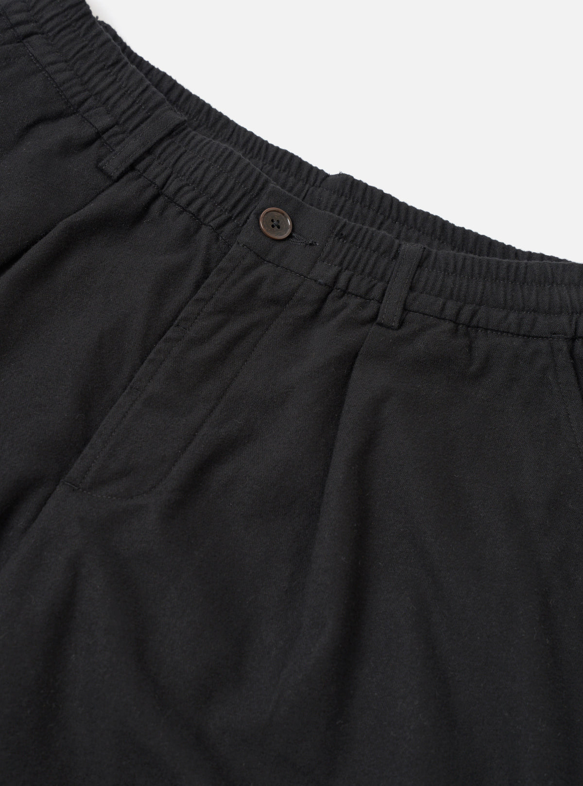 Close-up flat lay of black brushed moleskin pleated track pants on a white studio backdrop. The elasticated waistband with ruched gathering is visible at the top, alongside a single dark horn button fastening. A deep patch pocket sits at the front left, and soft fabric drape highlights the pleated construction below the waistband.