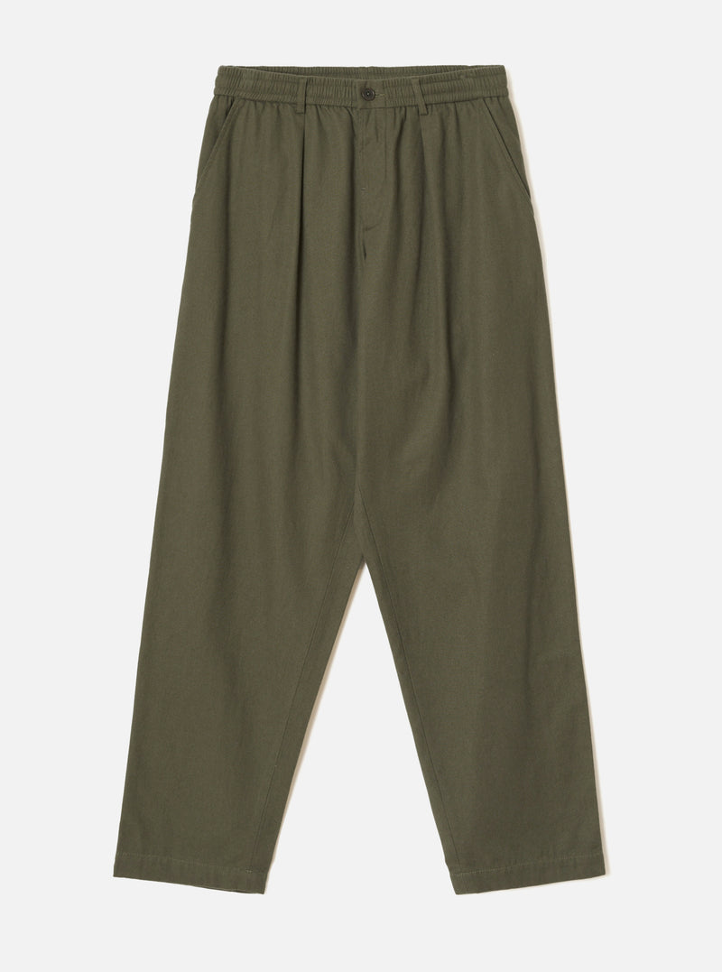 Olive brushed moleskin pleated track pants laid flat on a plain light grey studio backdrop. The elasticated waistband features a single dark button at the centre front. Pleats run from waist to thigh on each leg, with slanted side pockets. The wide leg tapers slightly toward the cropped hem.