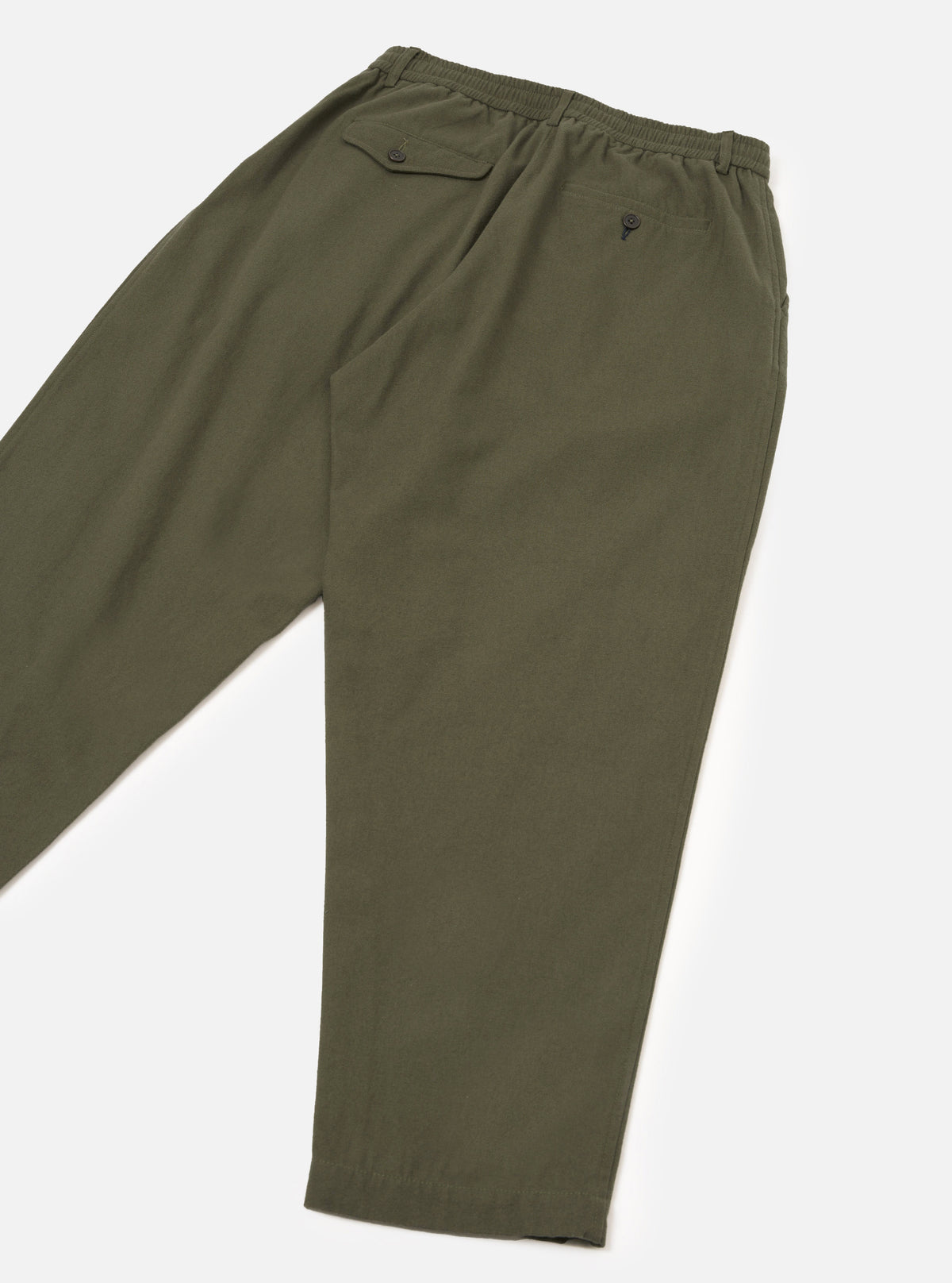 Olive brushed moleskin track pants laid flat on a white studio surface, angled view showing the back. Elasticated waistband with a buttoned flap pocket on the left hip and a single button-fastened welt pocket at centre back. Relaxed tapered leg in plain smooth olive fabric.
