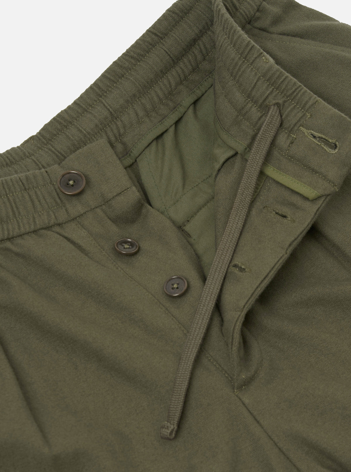 Close-up flat lay of olive brushed moleskin pleated track pants on a light grey backdrop. The elasticated waistband with ribbed texture is visible at the top, alongside a matching olive drawcord. The fly features three dark brown buttons. The lining is visible where the fly sits open.