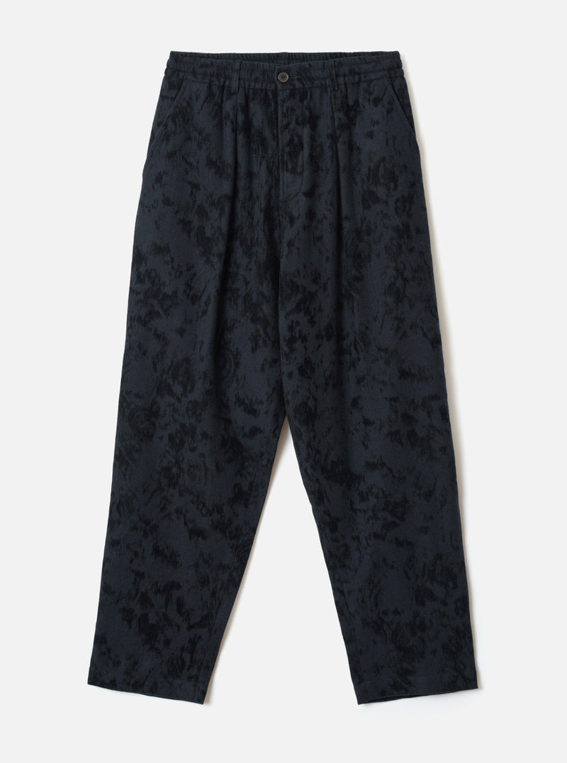 Black wool-mix track pants with an all-over abstract blurred print in dark navy and black tones. Features an elasticated waistband with a single centre button, twin pleats at the front, side slash pockets, and a relaxed tapered leg to an unfinished hem. Flat lay on a light grey studio backdrop.