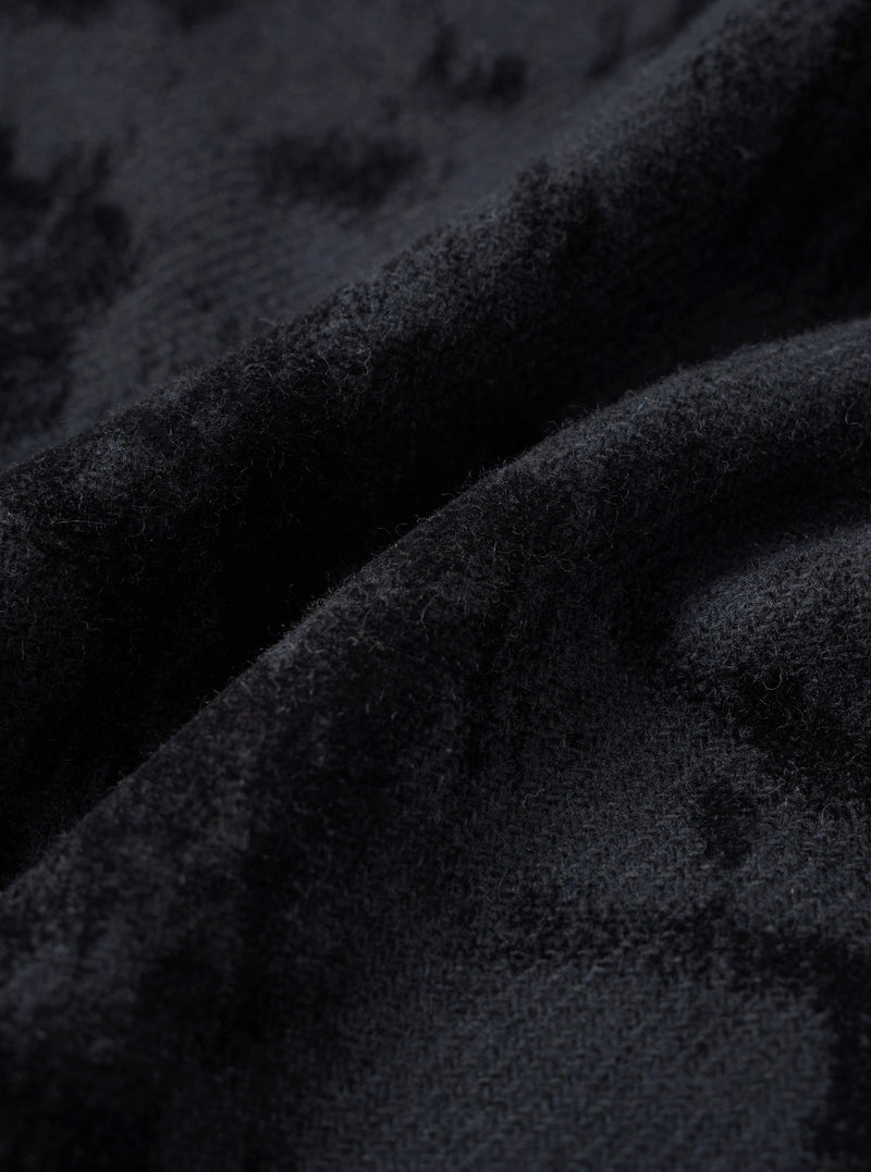 Close-up detail of the black wool mix fabric used in the pleated track pant. The surface has a dense, lightly boiled or brushed texture with fine fibres visible throughout. Deep folds in the fabric reveal soft drape and a matte finish with subtle tonal variation across the dark charcoal-black cloth.