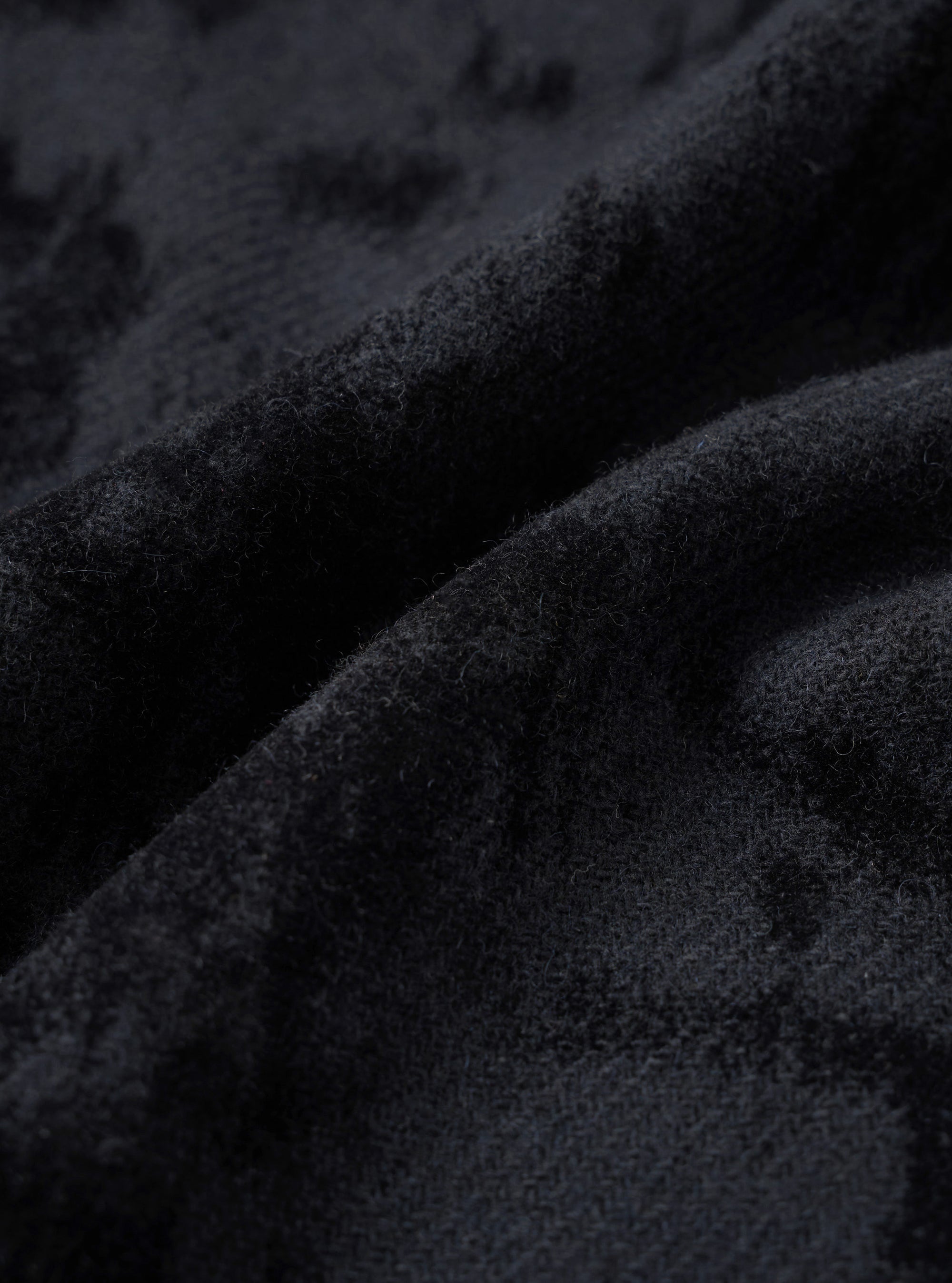 Close-up detail of the black wool mix fabric used in the pleated track pant. The surface has a dense, lightly boiled or brushed texture with fine fibres visible throughout. Deep folds in the fabric reveal soft drape and a matte finish with subtle tonal variation across the dark charcoal-black cloth.