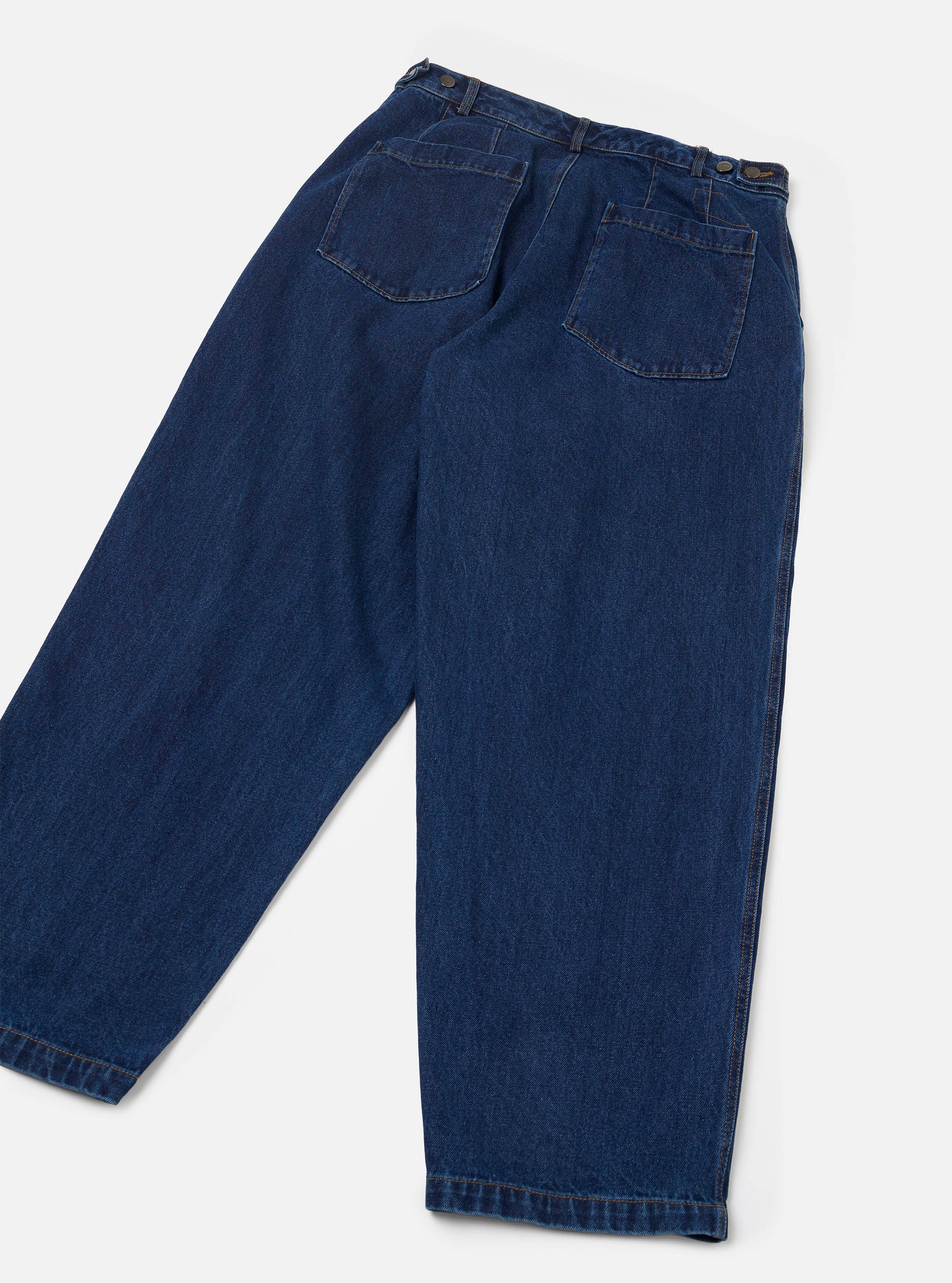 Flat lay of indigo work denim Dutch pants on a light grey seamless backdrop, shot from the back. Wide-leg silhouette with a high waist, two large patch pockets with topstitching, brass side-adjustor buttons at the waistband, and contrast orange stitching at the hems. Plain-woven mid-weight denim in deep indigo blue.