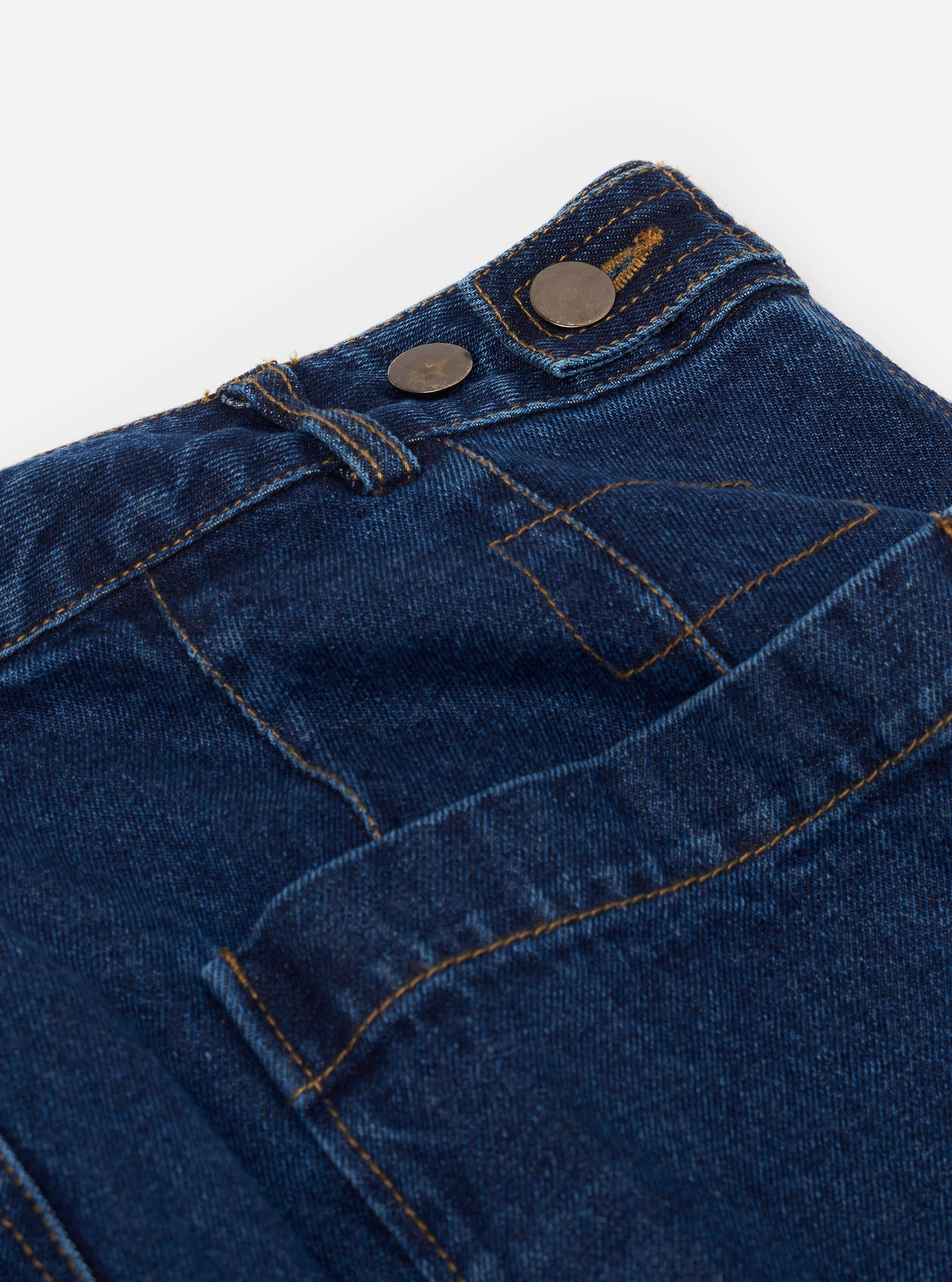 Close-up detail of the indigo work denim Dutch pant against a white studio backdrop. Two gunmetal snap buttons fasten the waistband extension. Gold topstitching outlines the waistband and a large angled front patch pocket. The dark indigo denim has a slightly worn texture.