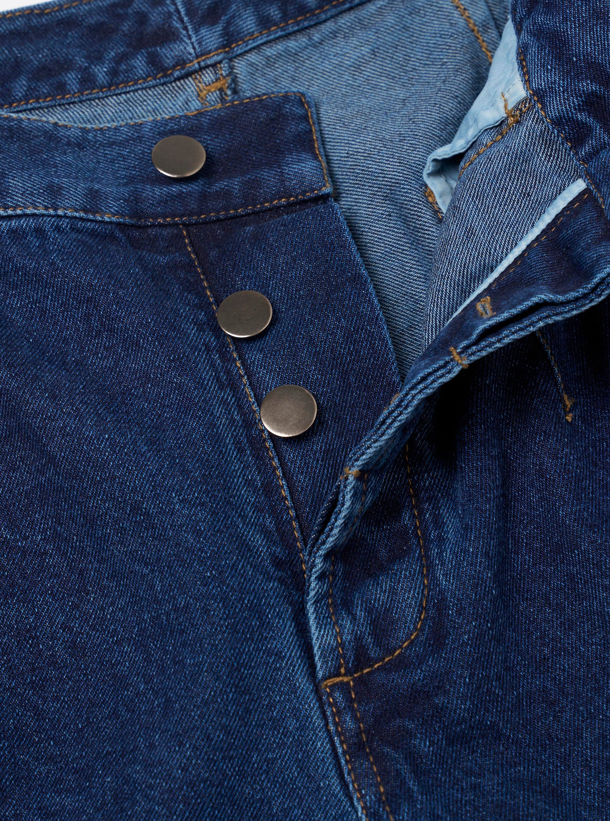 Close-up detail of the indigo work denim Dutch pant waistband and fly. Three gunmetal press-stud buttons run down the open fly placket, with the waistband folded back to reveal a light blue lining. Amber topstitching is visible throughout on the dark indigo twill fabric. Plain white studio backdrop.