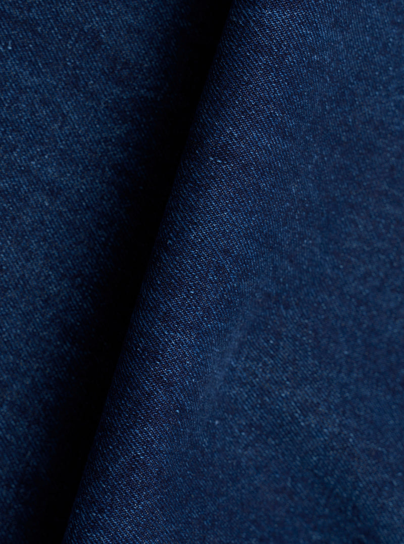 Close-up of the indigo denim fabric used in the Work Dutch Pant. The woven textile shows a fine, slightly slubby texture with tonal variation across the deep navy-indigo surface. The fabric is folded, creating a shadow crease that highlights the cloth weight and weave structure.