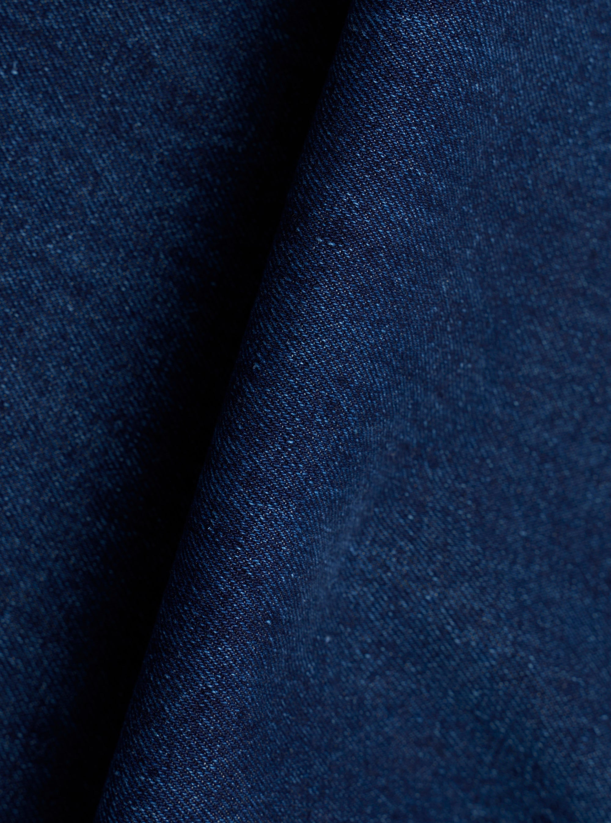 Close-up of the indigo denim fabric used in the Work Dutch Pant. The woven textile shows a fine, slightly slubby texture with tonal variation across the deep navy-indigo surface. The fabric is folded, creating a shadow crease that highlights the cloth weight and weave structure.