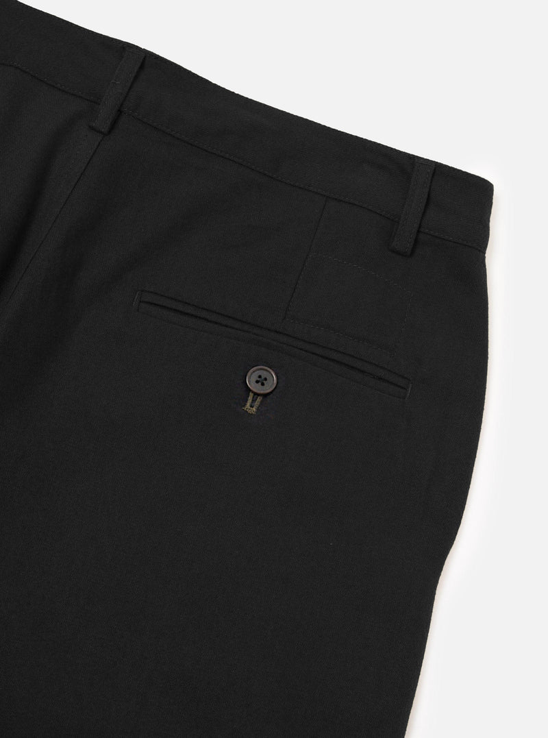 Close-up flat lay of black brushed moleskin military chinos on a white studio surface, showing the rear waistband with two belt loops, a single welt back pocket fastened with a dark four-hole button and contrast bartack stitching. The fabric has a smooth, fine-brushed texture.