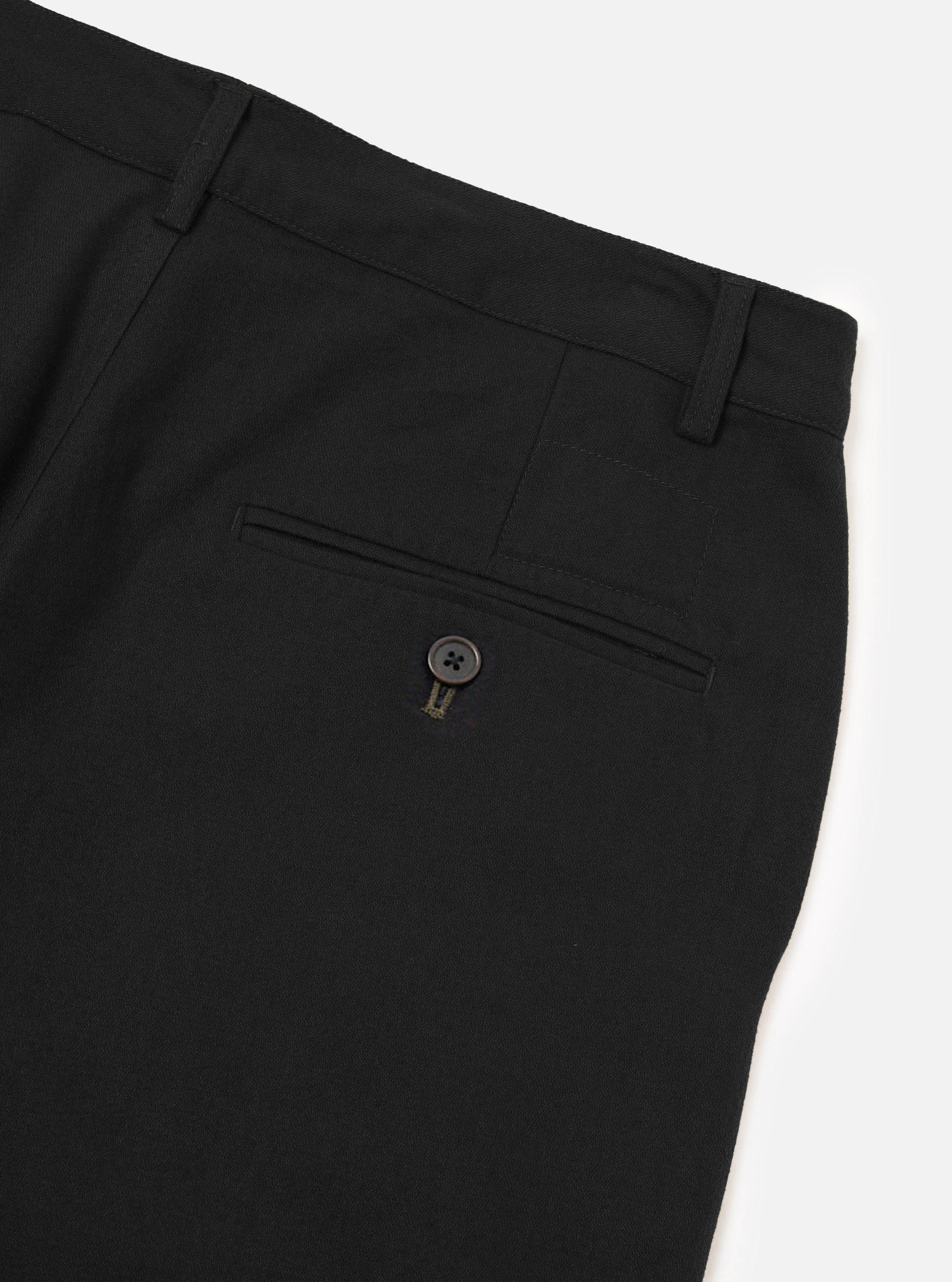 Close-up flat lay of black brushed moleskin military chinos on a white studio surface, showing the rear waistband with two belt loops, a single welt back pocket fastened with a dark four-hole button and contrast bartack stitching. The fabric has a smooth, fine-brushed texture.