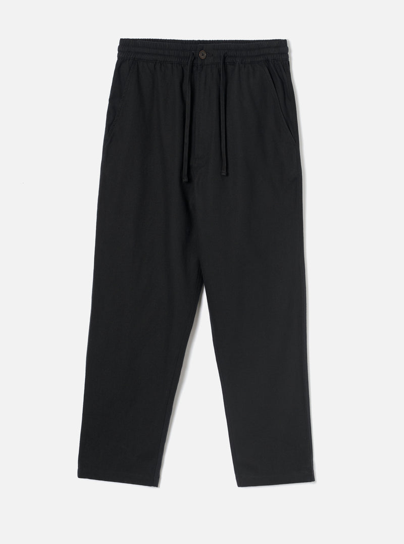 Black brushed moleskin hi water trousers laid flat on a plain light grey studio backdrop. The elasticated waistband features a dark drawcord and a single dark button at the centre front. Slanted side pockets sit at the hips. The relaxed leg tapers to a cropped, unfinished hem. Fabric has a soft, brushed texture throughout.