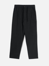 Black brushed moleskin hi water trousers laid flat on a plain light grey studio backdrop. The elasticated waistband features a dark drawcord and a single dark button at the centre front. Slanted side pockets sit at the hips. The relaxed leg tapers to a cropped, unfinished hem. Fabric has a soft, brushed texture throughout.