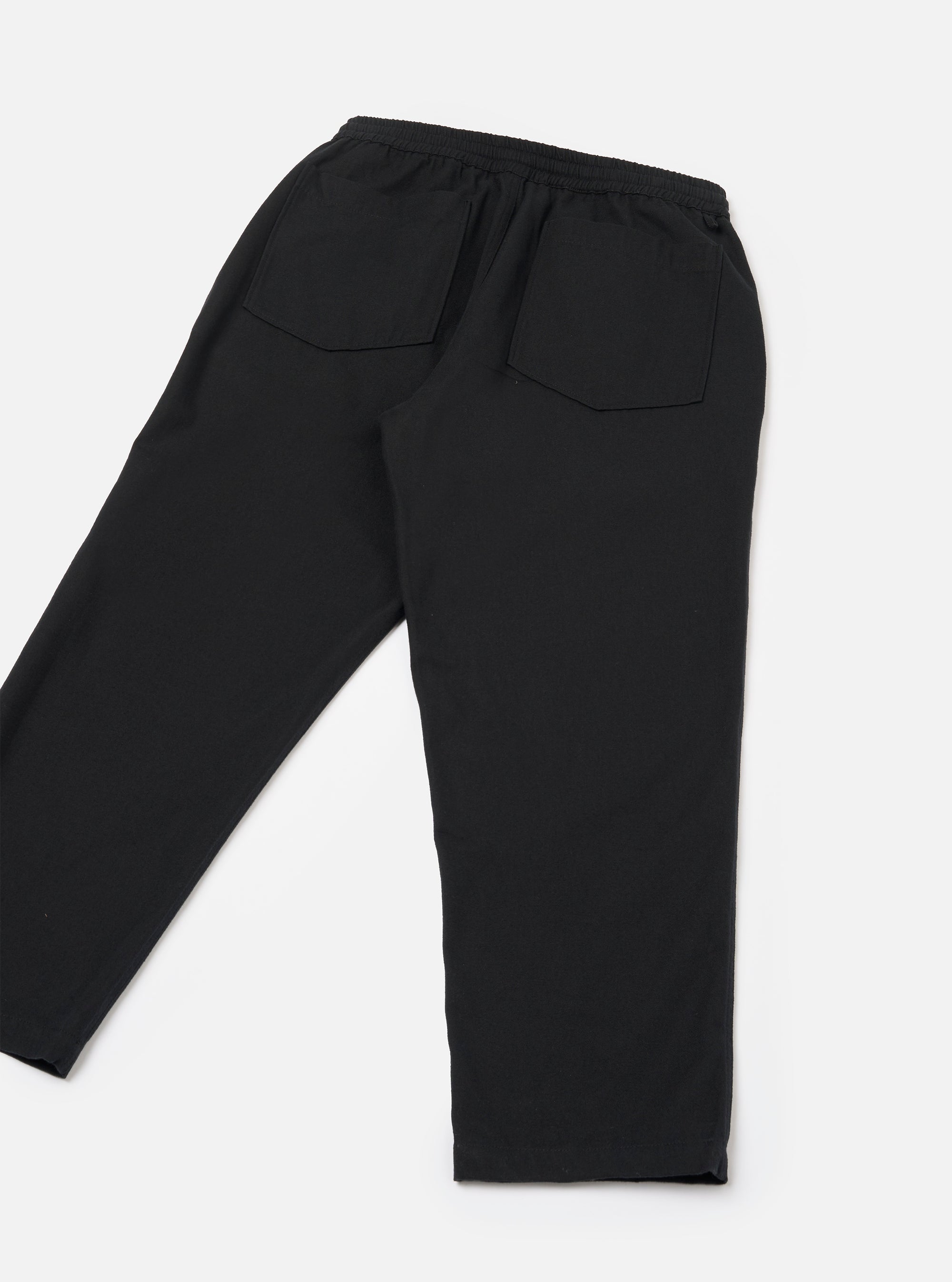 Black brushed moleskin hi-water trousers laid flat on a white studio surface, shot from the back. The elasticated waistband with a visible drawcord is prominent at the top. Two large patch pockets sit on the rear. The tapered legs end in a cropped, unfinished hem. Plain, smooth fabric with a subtle matte finish.