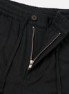 Close-up flat lay of black brushed moleskin trousers on a white studio backdrop. The shot focuses on the waistband and fly area, showing a ribbed elasticated waistband, an open silver metal zip fly, a single dark brown button fastening, and a zip-entry side pocket on the right.