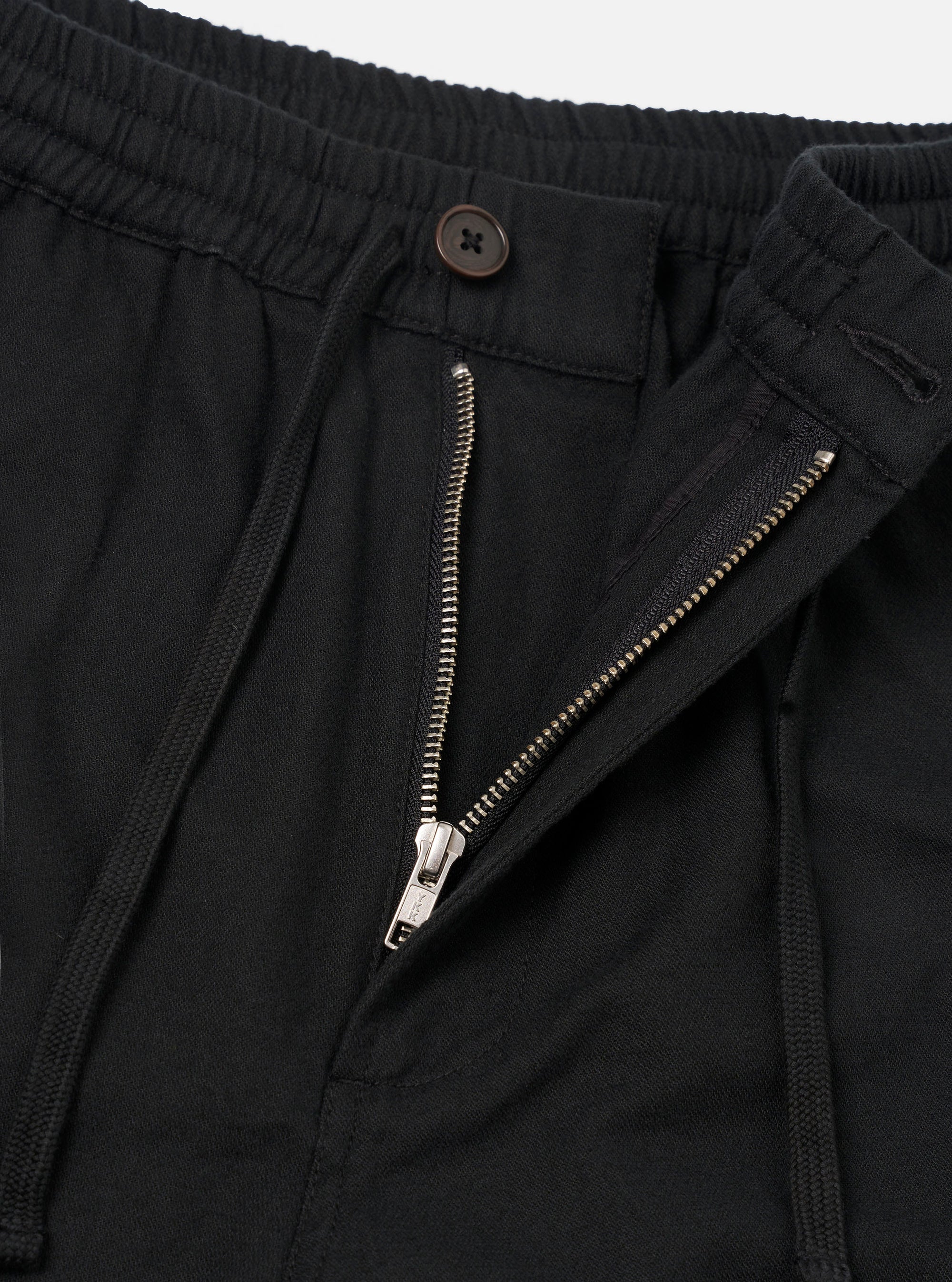 Close-up flat lay of black brushed moleskin trousers on a white studio backdrop. The shot focuses on the waistband and fly area, showing a ribbed elasticated waistband, an open silver metal zip fly, a single dark brown button fastening, and a zip-entry side pocket on the right.