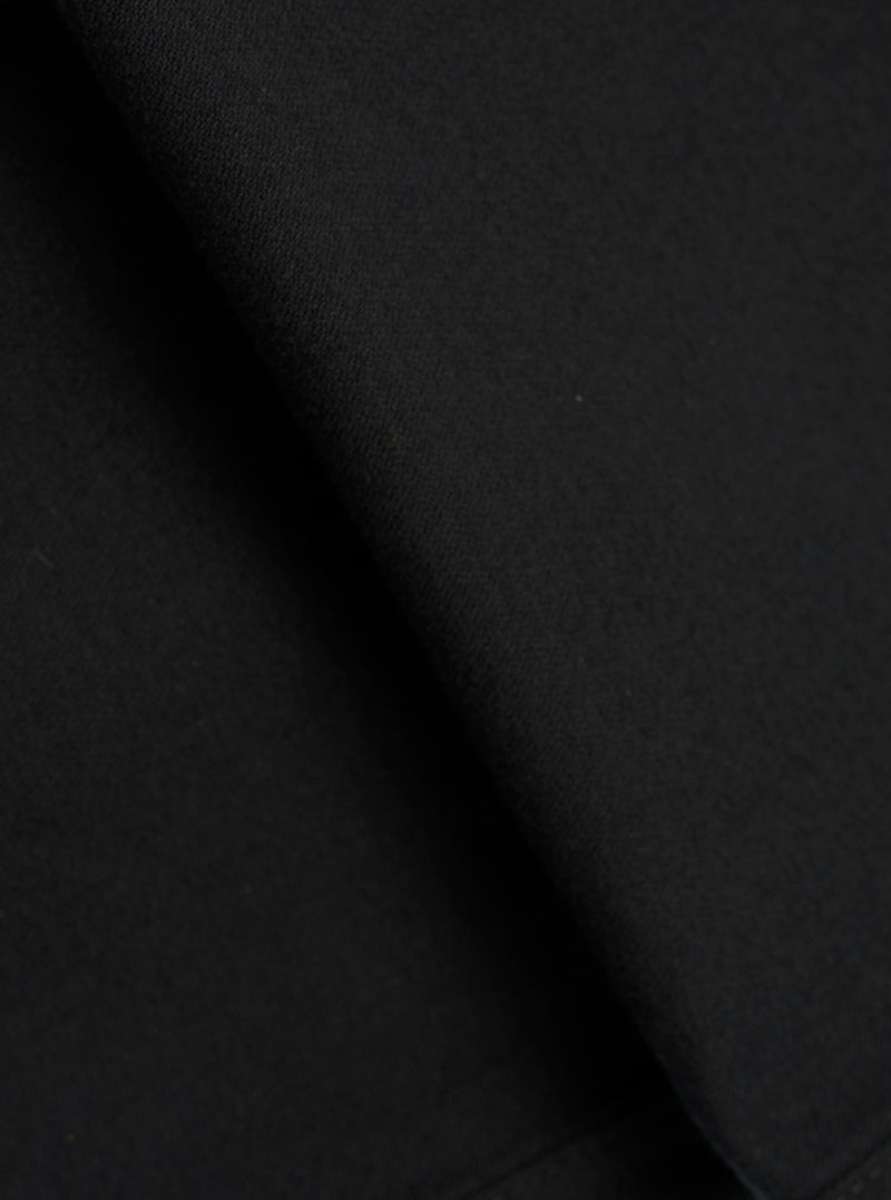 Close-up fabric detail of black brushed moleskin trousers, showing the dense, soft-napped texture of the cloth. The tight knit weave is visible in the upper corner, contrasting with the smooth brushed surface. Deep folds create areas of shadow across the matte black fabric.