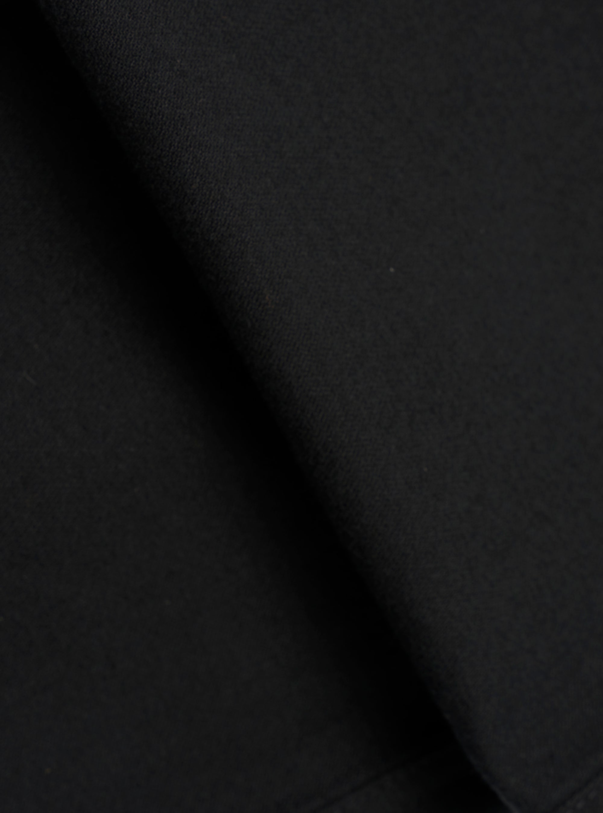 Close-up fabric detail of black brushed moleskin trousers, showing the dense, soft-napped texture of the cloth. The tight knit weave is visible in the upper corner, contrasting with the smooth brushed surface. Deep folds create areas of shadow across the matte black fabric.