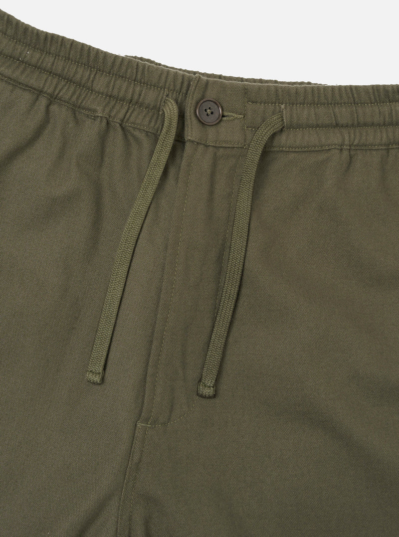 Close-up of the waistband on olive brushed moleskin hi water trousers. The elasticated waistband features a matching fabric-covered section, a dark four-hole button, and a woven drawcord threaded through two eyelets hanging loose. Plain white studio backdrop.