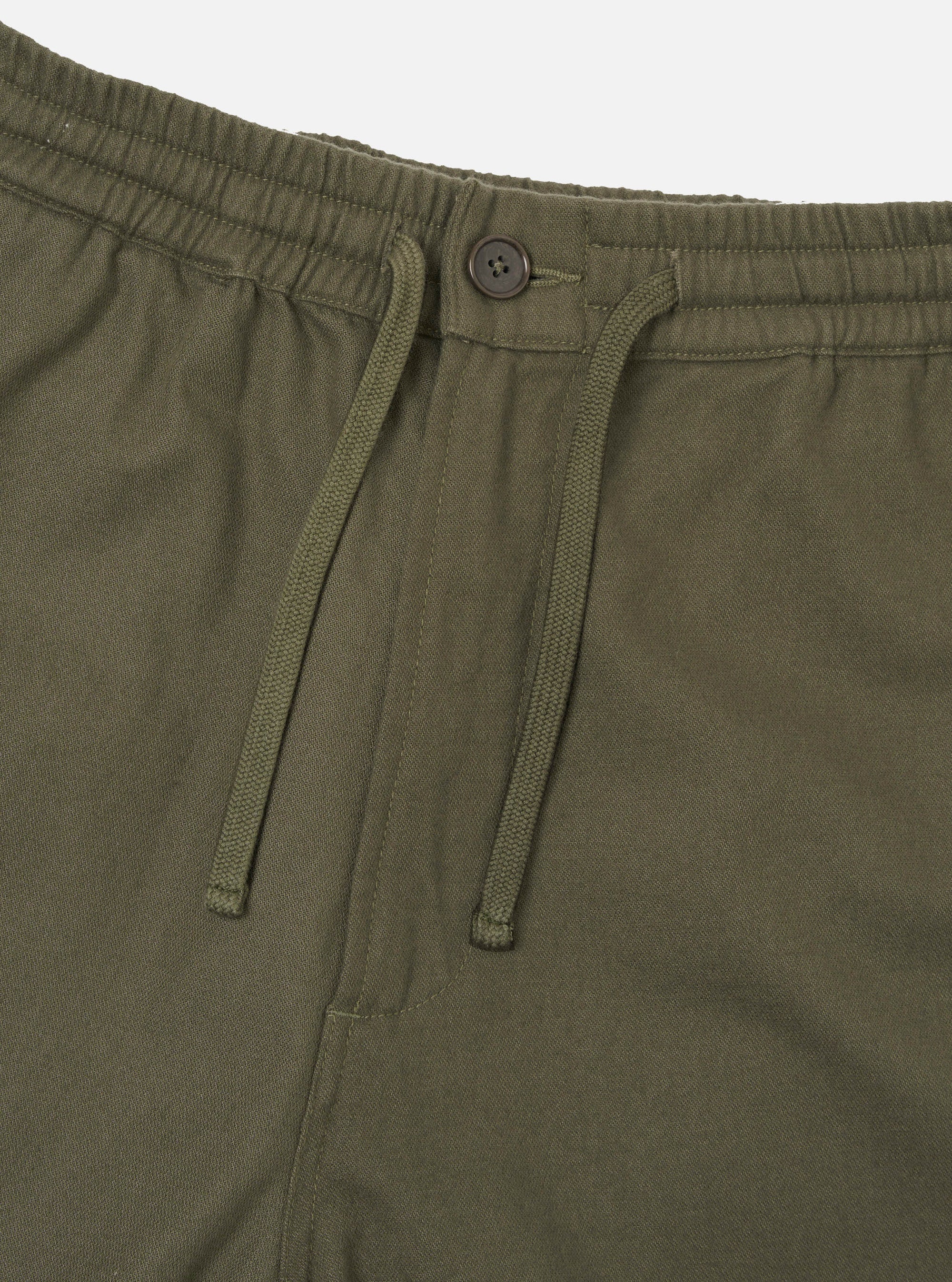 Close-up of the waistband on olive brushed moleskin hi water trousers. The elasticated waistband features a matching fabric-covered section, a dark four-hole button, and a woven drawcord threaded through two eyelets hanging loose. Plain white studio backdrop.
