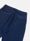 Flat lay close-up of indigo work denim super chinos on a light grey studio backdrop. The rear waistband, two buttoned welt back pockets with brass button closures, and topstitching in gold thread are clearly visible. The wide trouser legs are cropped just above the hem.