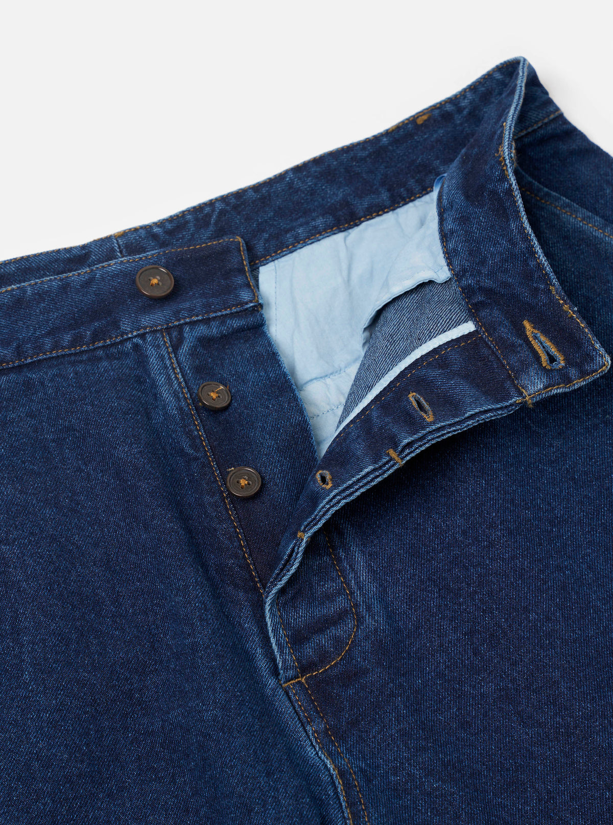 Close-up flat lay of indigo work denim super chinos on a light grey studio backdrop. The shot focuses on the open button fly, showing four dark metal buttons and four corresponding buttonholes on the folded fly flap. Yellow-gold contrast stitching runs throughout. The pale blue fabric lining is visible inside the waistband.