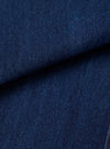 Close-up fabric shot of indigo work denim, showing a deep navy blue twill weave with a tight, dense texture. The fabric is folded to reveal depth and weight. A small section of gold-tan stitching is visible at the bottom right corner. No background is visible.