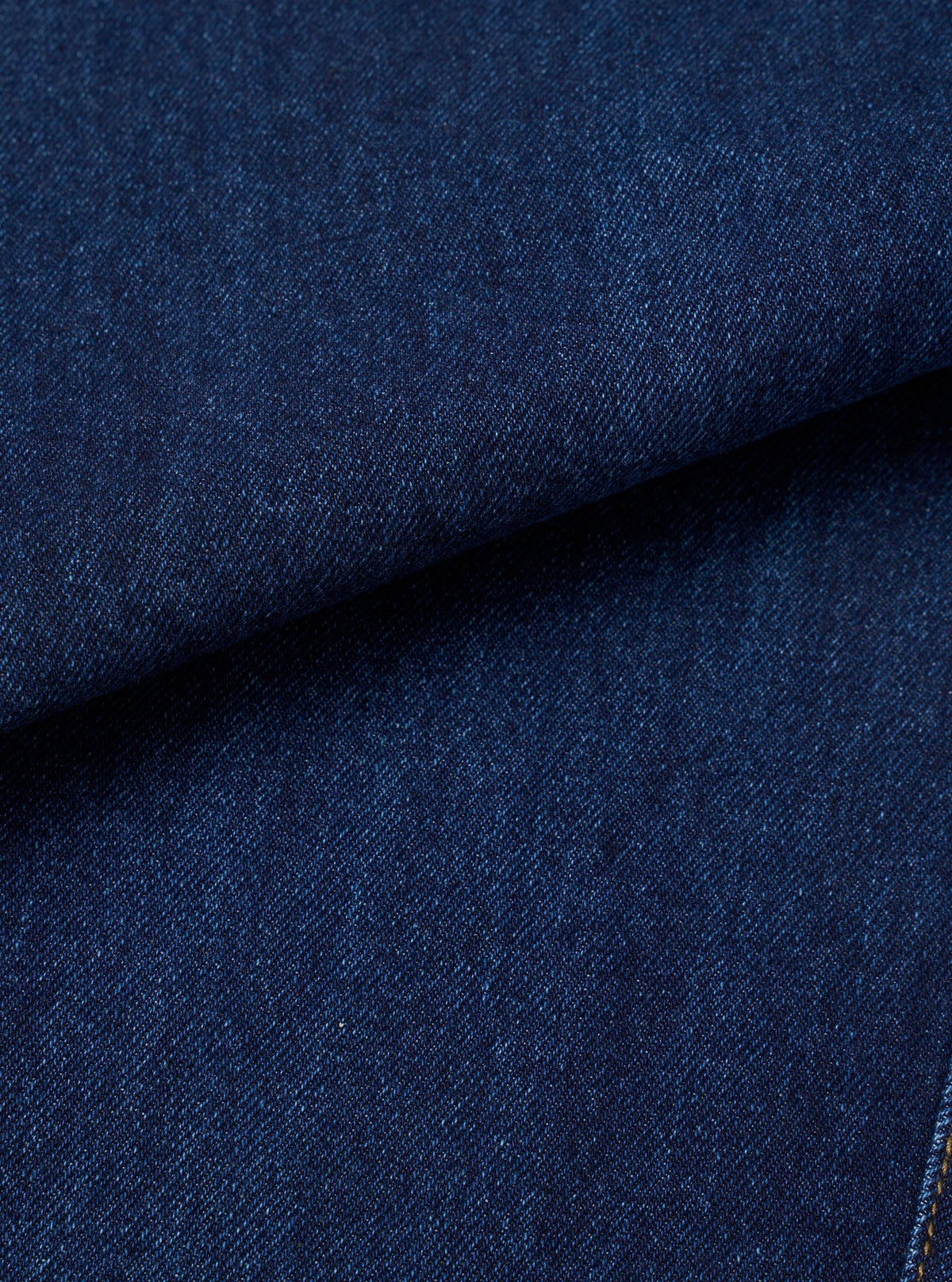 Close-up fabric shot of indigo work denim, showing a deep navy blue twill weave with a tight, dense texture. The fabric is folded to reveal depth and weight. A small section of gold-tan stitching is visible at the bottom right corner. No background is visible.