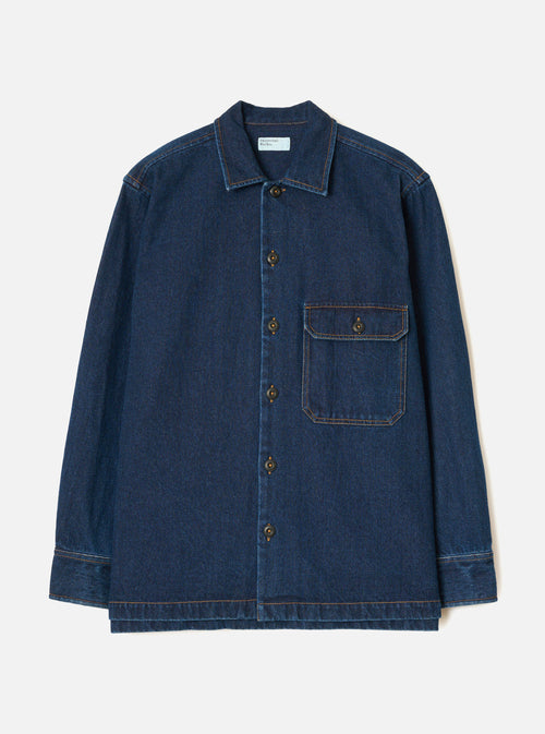 Indigo denim fatigue shirt on a plain light grey backdrop, no model. Features a camp collar, five bronze buttons down the placket, and a single chest patch pocket with a buttoned flap. Contrast orange topstitching throughout. Long sleeves with wide buttoned cuffs and a straight, boxy hem. Universal Works woven label at the chest.