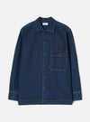 Indigo denim fatigue shirt on a plain light grey backdrop, no model. Features a camp collar, five bronze buttons down the placket, and a single chest patch pocket with a buttoned flap. Contrast orange topstitching throughout. Long sleeves with wide buttoned cuffs and a straight, boxy hem. Universal Works woven label at the chest.
