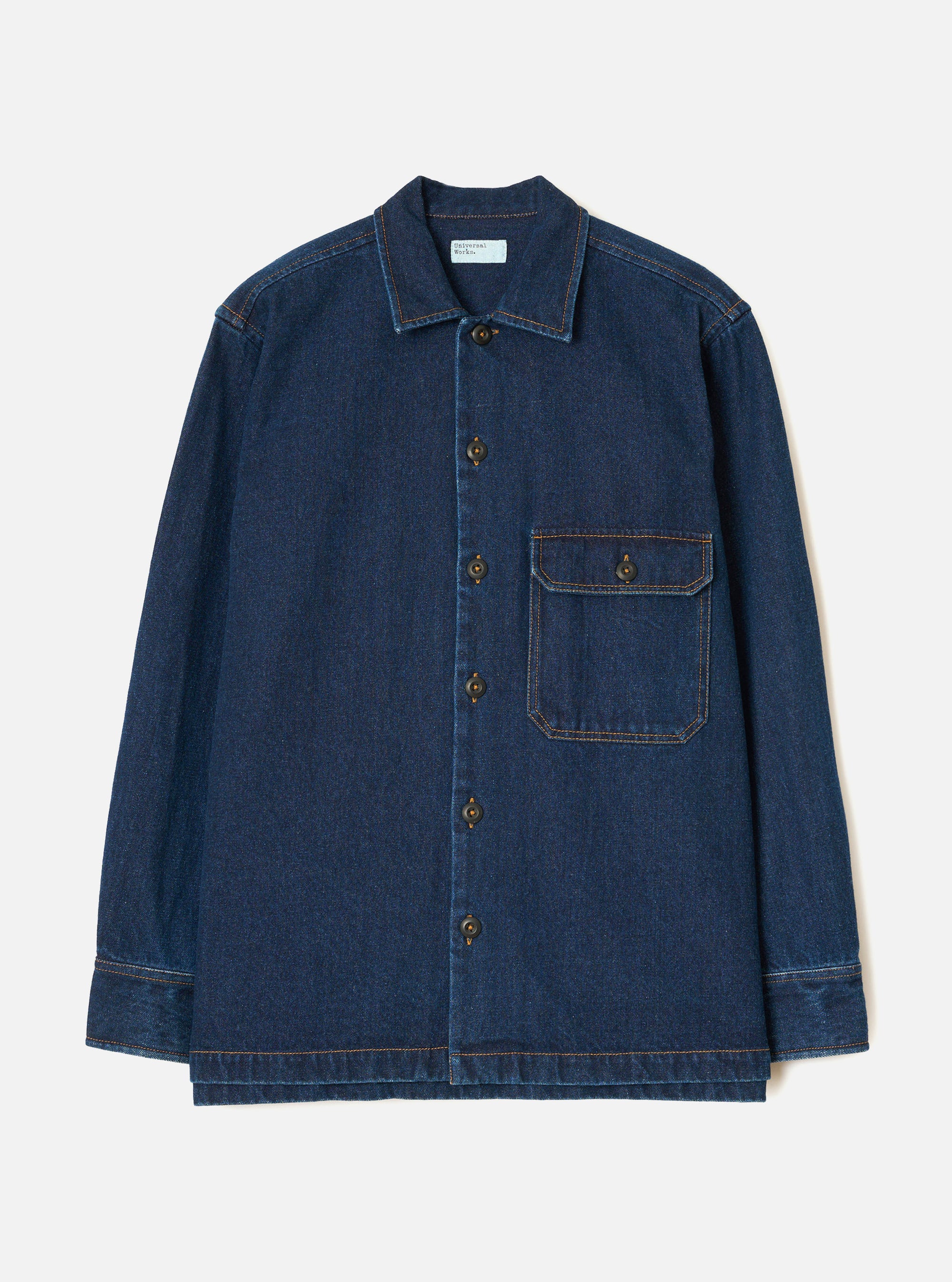 Indigo denim fatigue shirt on a plain light grey backdrop, no model. Features a camp collar, five bronze buttons down the placket, and a single chest patch pocket with a buttoned flap. Contrast orange topstitching throughout. Long sleeves with wide buttoned cuffs and a straight, boxy hem. Universal Works woven label at the chest.