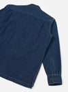 Rear flat-lay of an indigo denim fatigue shirt on a light grey seamless backdrop. The back yoke, orange contrast topstitching at seams and shoulder, a rear patch pocket outline at the hem, and a single long sleeve are visible. The cuff features a buttoned placket with two dark buttons.