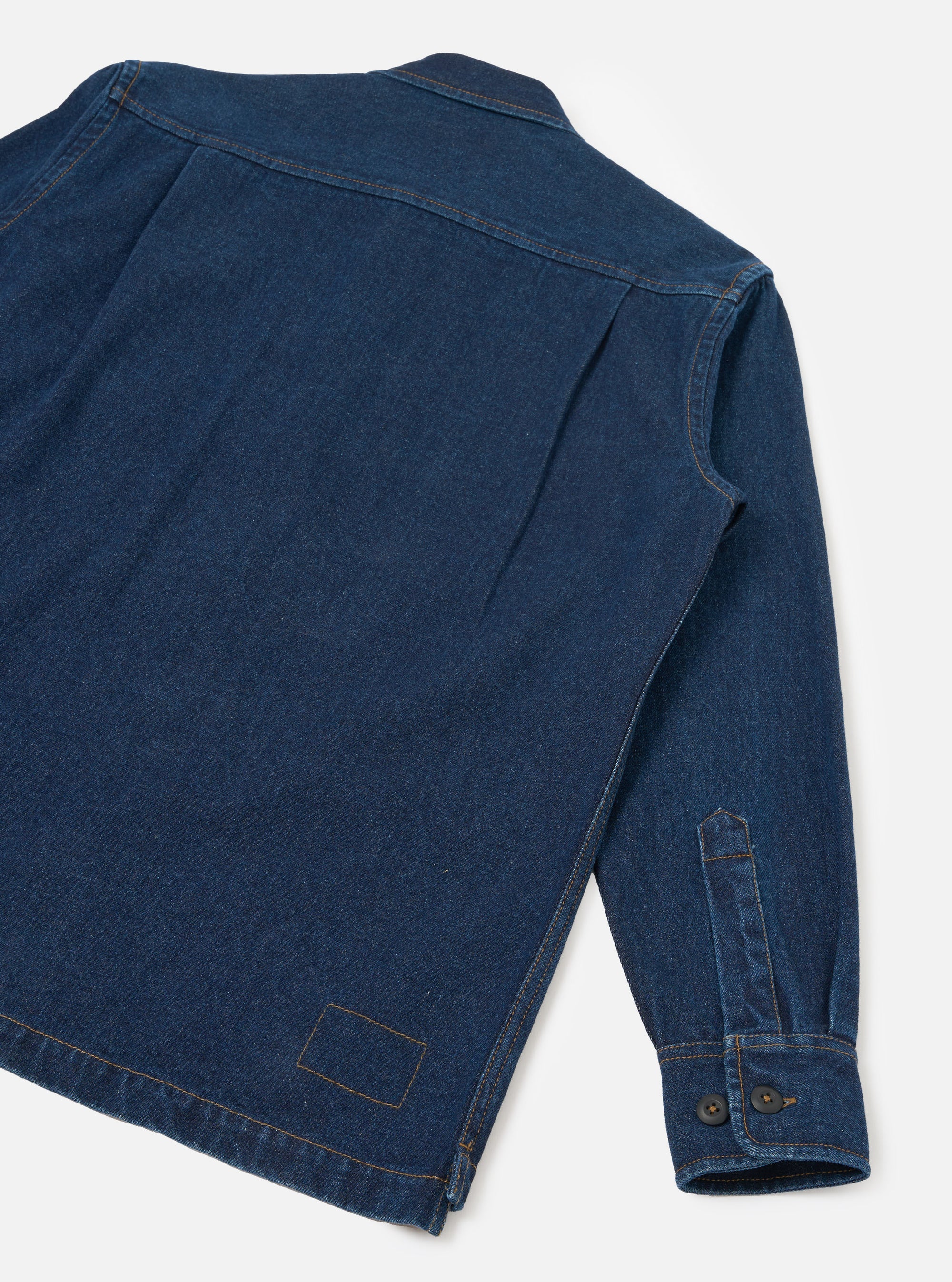 Rear flat-lay of an indigo denim fatigue shirt on a light grey seamless backdrop. The back yoke, orange contrast topstitching at seams and shoulder, a rear patch pocket outline at the hem, and a single long sleeve are visible. The cuff features a buttoned placket with two dark buttons.