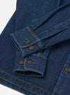 Close-up flat lay of an indigo denim fatigue shirt on a white surface, focusing on the cuffs and front placket. Two buttoned barrel cuffs feature dark horn-style buttons and contrast amber topstitching throughout. A button-through placket with matching dark buttons is visible at the lower right.
