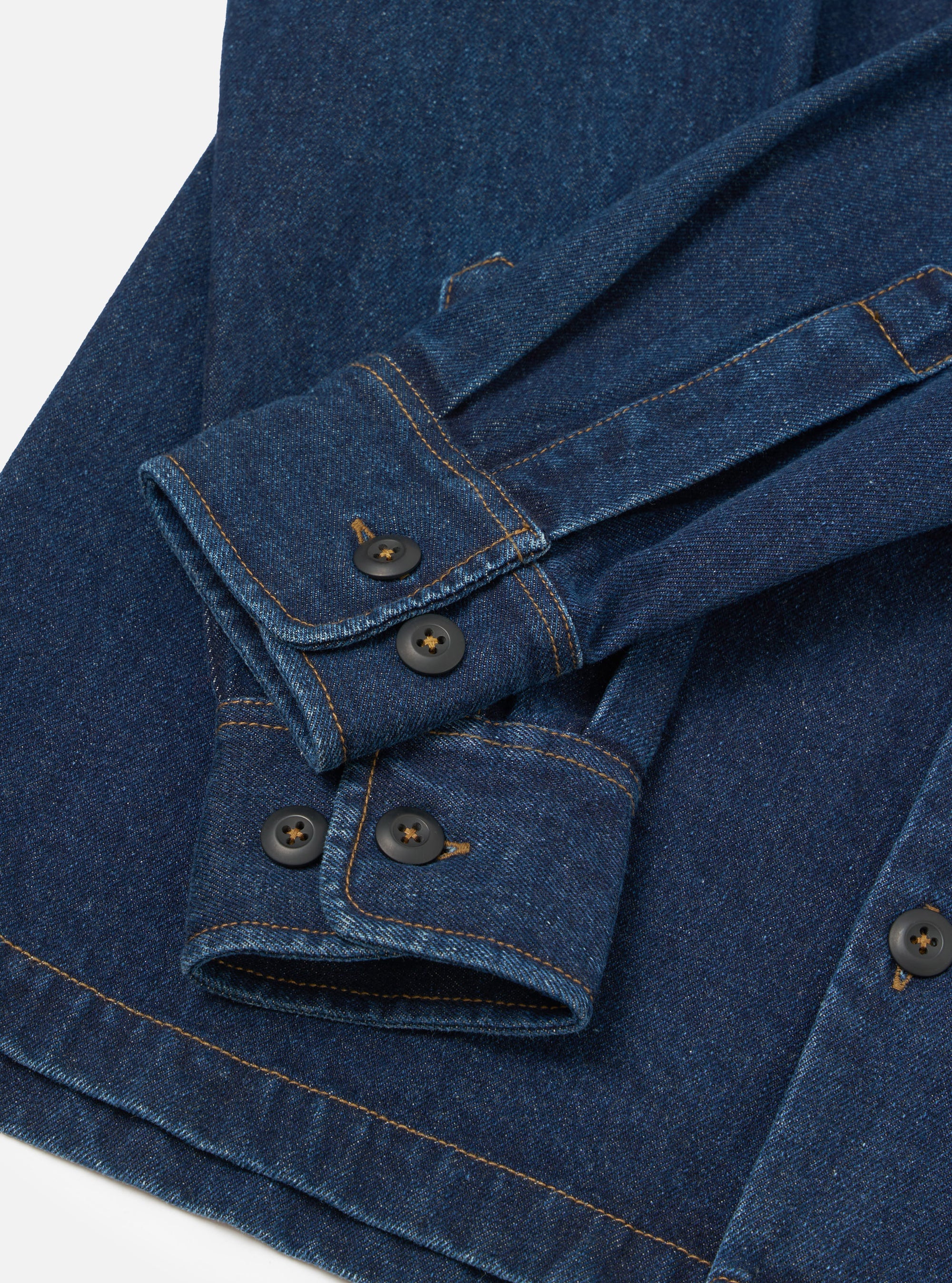 Close-up flat lay of an indigo denim fatigue shirt on a white surface, focusing on the cuffs and front placket. Two buttoned barrel cuffs feature dark horn-style buttons and contrast amber topstitching throughout. A button-through placket with matching dark buttons is visible at the lower right.
