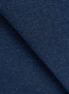 Close-up of indigo denim fabric folded diagonally across the frame, showing the twill weave texture with flecks of blue and dark thread throughout. The dense, structured fabric has a matte finish with subtle variation in tone across the surface.