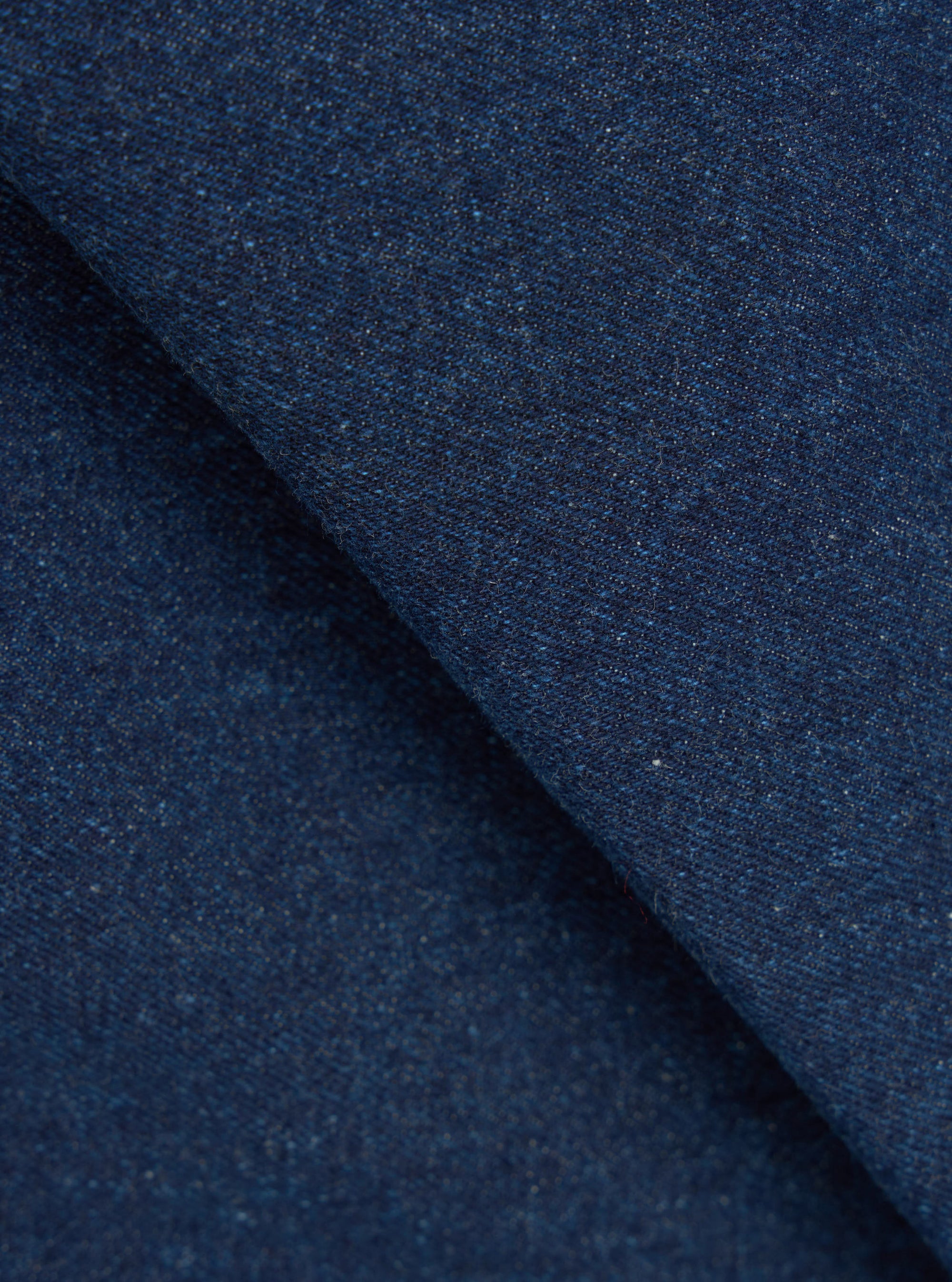 Close-up of indigo denim fabric folded diagonally across the frame, showing the twill weave texture with flecks of blue and dark thread throughout. The dense, structured fabric has a matte finish with subtle variation in tone across the surface.