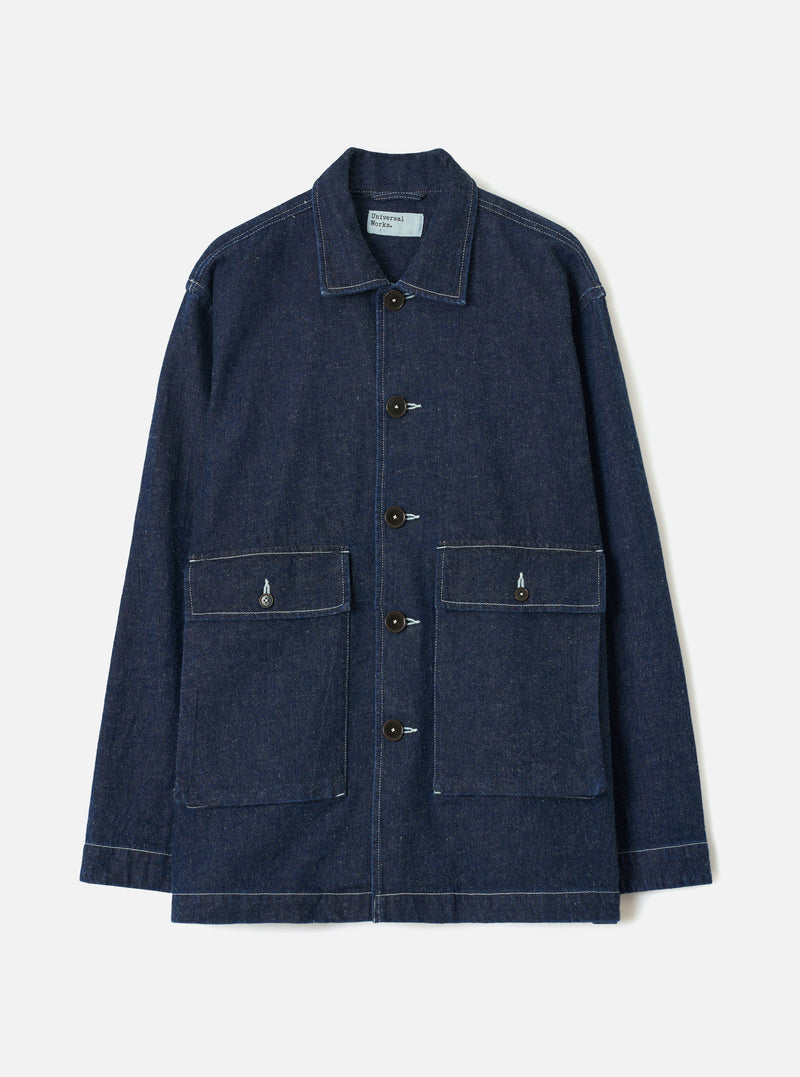Indigo denim chore jacket laid flat on a light grey studio backdrop. Features a point collar, five dark buttons down the placket, two large patch pockets with button flaps at the chest, and a lower patch pocket. Contrast white topstitching throughout. Universal Works woven label visible at the collar.