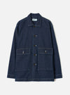 Indigo denim chore jacket laid flat on a light grey studio backdrop. Features a point collar, five dark buttons down the placket, two large patch pockets with button flaps at the chest, and a lower patch pocket. Contrast white topstitching throughout. Universal Works woven label visible at the collar.