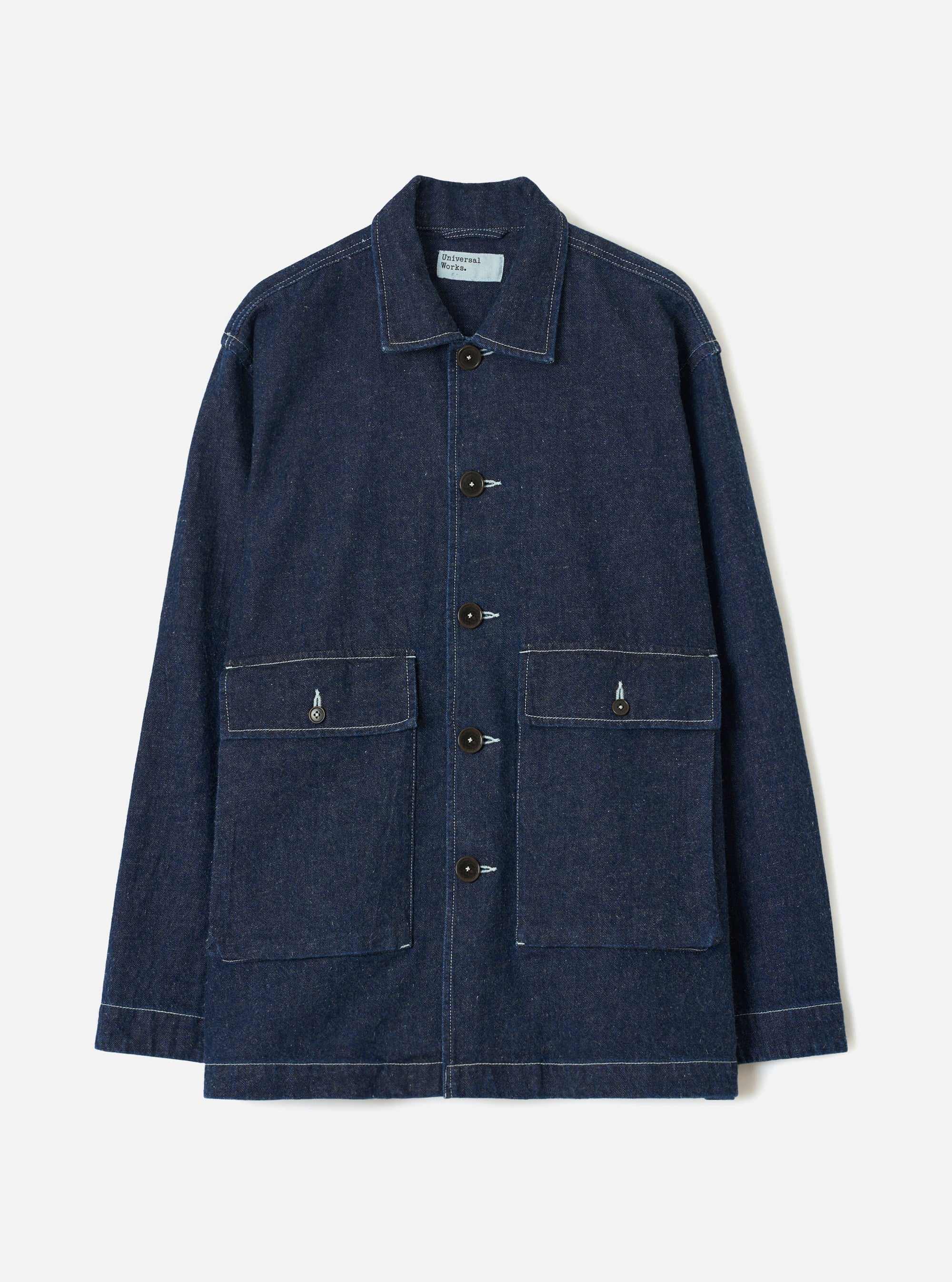Indigo denim chore jacket laid flat on a light grey studio backdrop. Features a point collar, five dark buttons down the placket, two large patch pockets with button flaps at the chest, and a lower patch pocket. Contrast white topstitching throughout. Universal Works woven label visible at the collar.