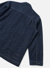 Rear three-quarter flat lay of an indigo denim troopers jacket on a white studio backdrop. Contrast white topstitching runs along the yoke, side seams, and hem. The cuff features two dark buttons. A stitched rectangular patch detail sits at the lower back hem.